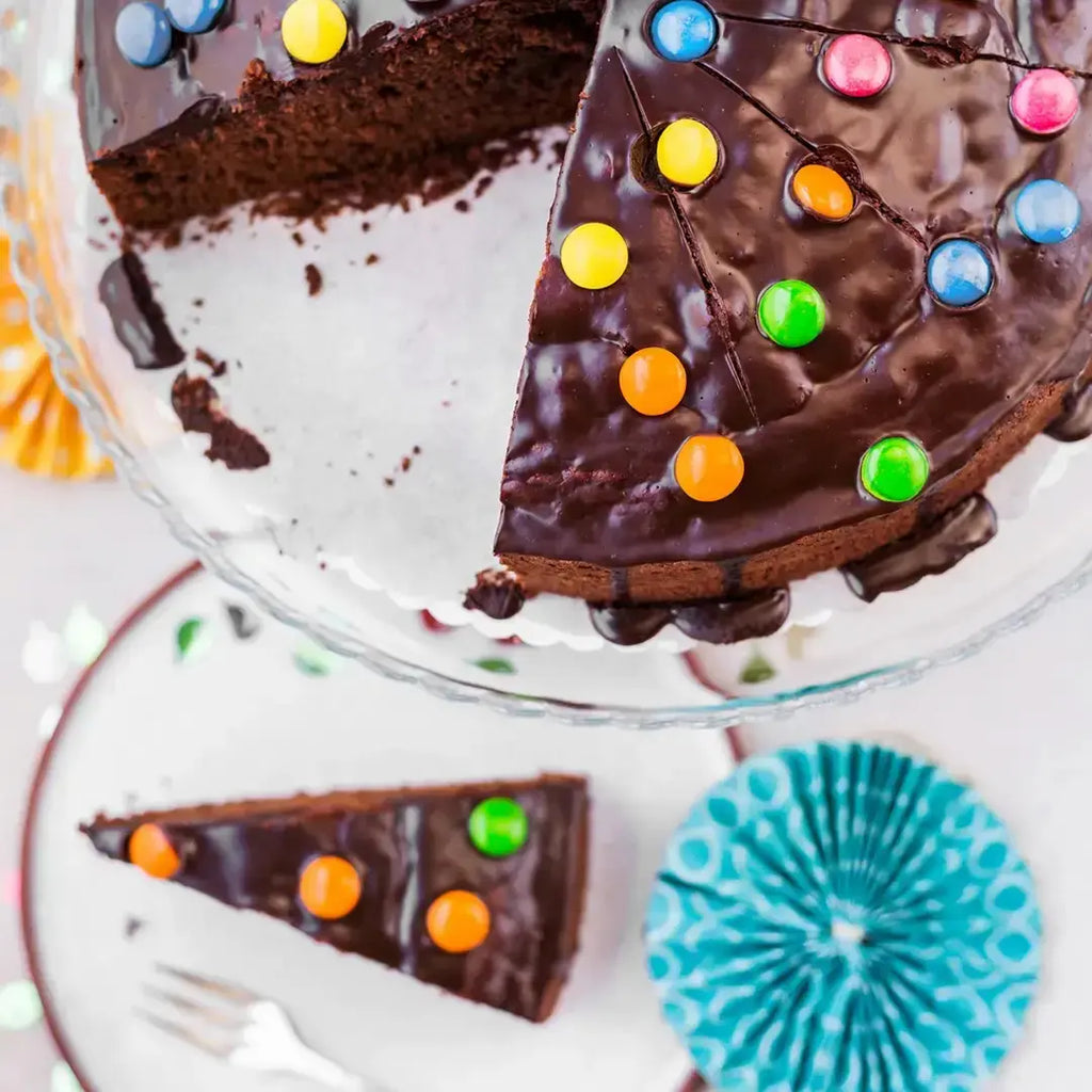 angeschnittener bunter Schokokuchen für Kinder von Oma Anna von Kuchentratsch auf Kuchenplatte und Teller, dekoriert mit Schokolinsen